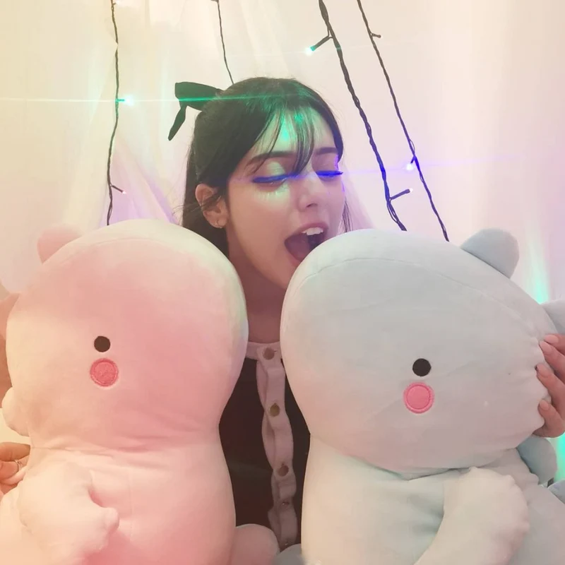 Playful young woman joking around with soft pink and light blue dinosaur plush toys, cozy room with fairy string lights, goofy cute stuffed animal friends